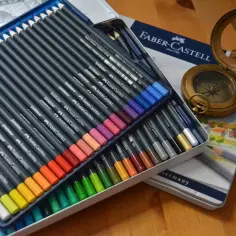 Zestaw kredek Faber-Castell Goldfaber w metalowym pudełku, obok kompas i zegar słoneczny. Różne rodzaje kredek czekają na artystę.