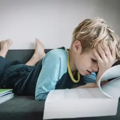 Chłopiec z ręką na czole, sfrustrowany nauką. Stos zeszytów obok. To trudny ii etap edukacyjny.