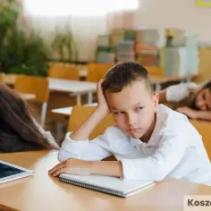 Zmęczony chłopiec z ręką na głowie siedzi przy ławce, jakby uczył się w szkole z czasów pruskiego systemu edukacji. Obok śpi koleżanka.