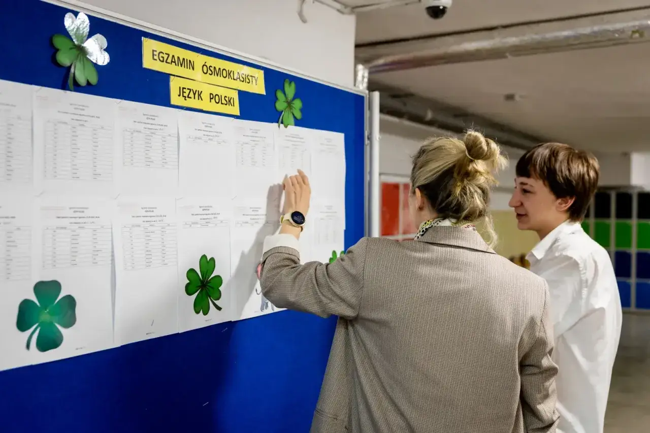 Wyniki egzaminu ósmoklasisty z języka polskiego. Czy oceny są ważne? Dwie osoby analizują tablicę z wynikami, ozdobioną koniczynami.