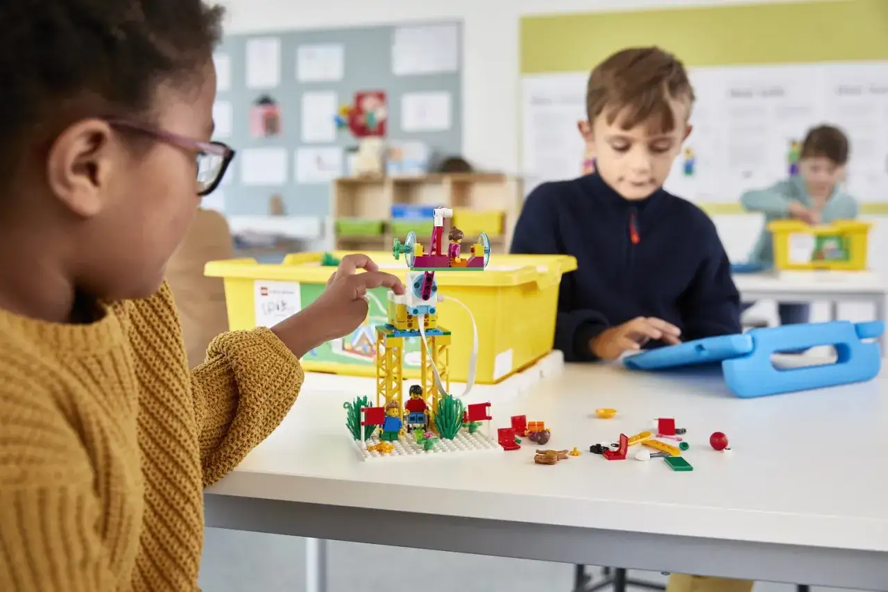 Dzieci budują z klocków podczas zajęć pozalekcyjnych. Dziewczynka z okularami i chłopiec z tabletem skupieni na tworzeniu.