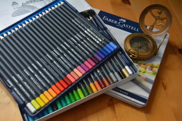 Zestaw kredek Faber-Castell Goldfaber w metalowym pudełku, obok kompas i zegar słoneczny. Różne rodzaje kredek czekają na artystę.